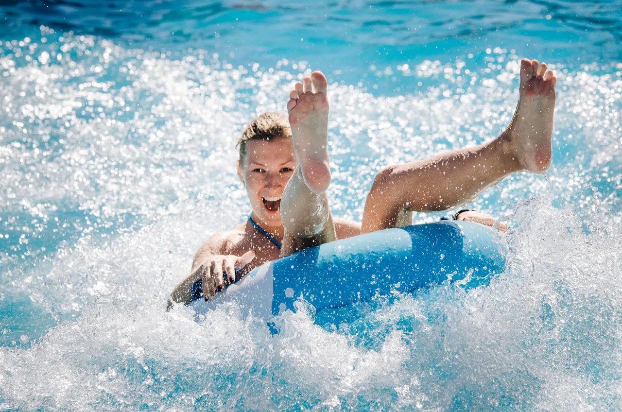 Child on a blue inflatable float splashing and playing in a wave pool with sparkling blue water.