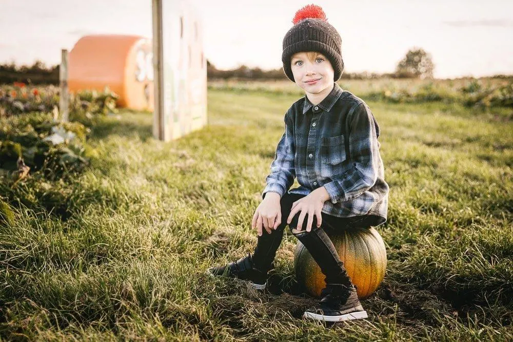 Child sitting on a pumpkin in a pumpkin patch field with farm buildings in the background.