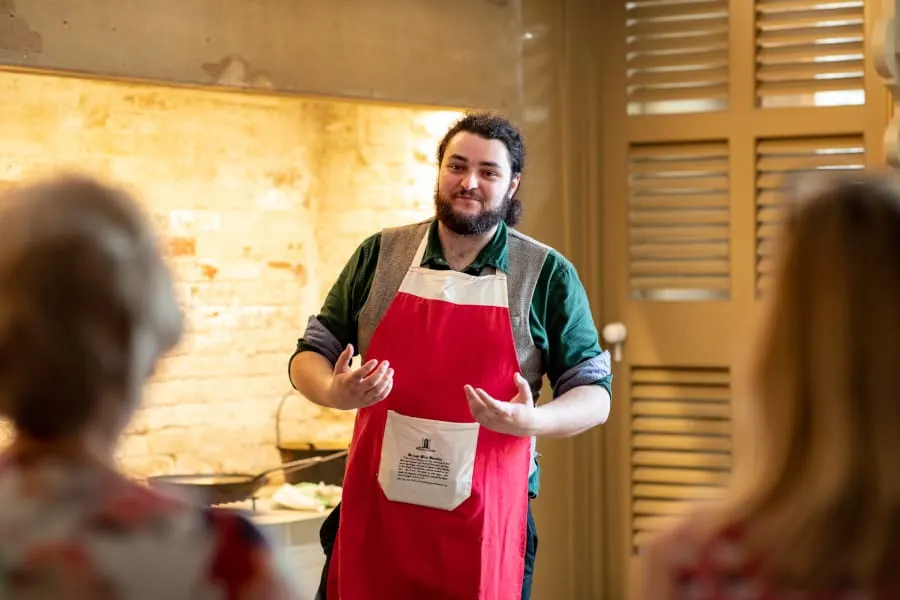Man wearing red apron speaking to visitors inside a historic stone room with wooden shutters.