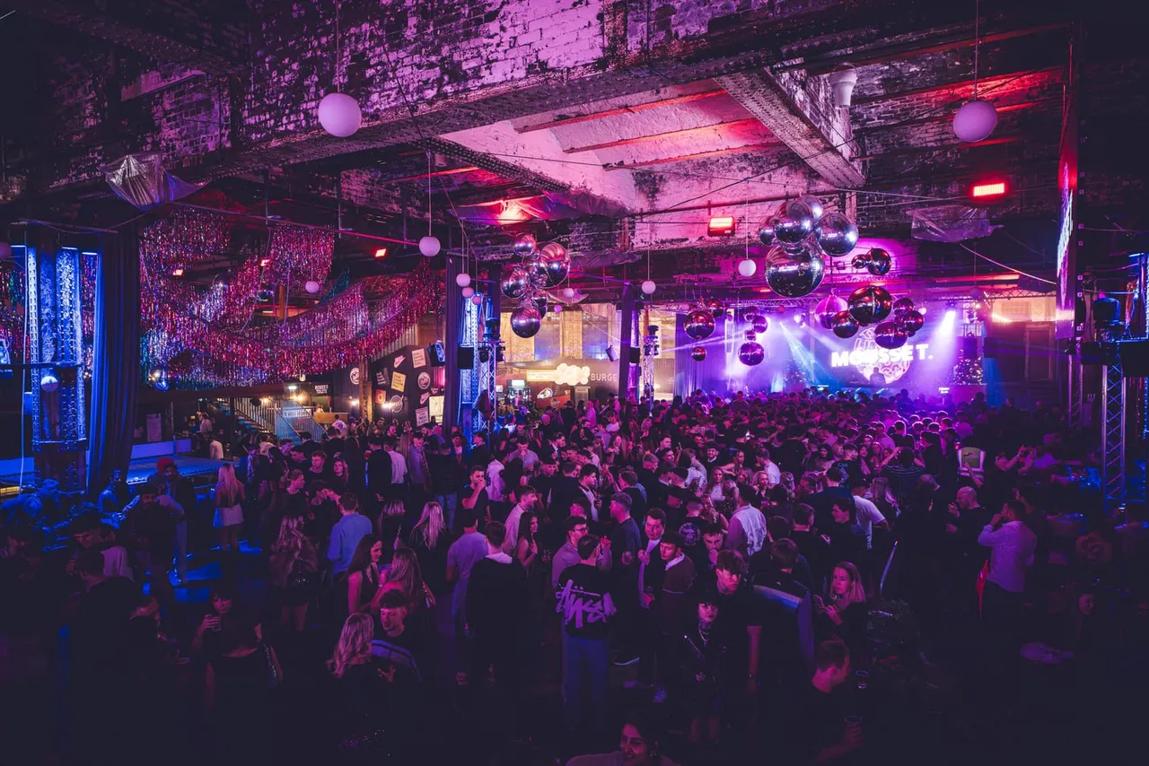 Crowded nightclub dance floor lit by pink and purple neon lights with disco balls and hanging decorations overhead.