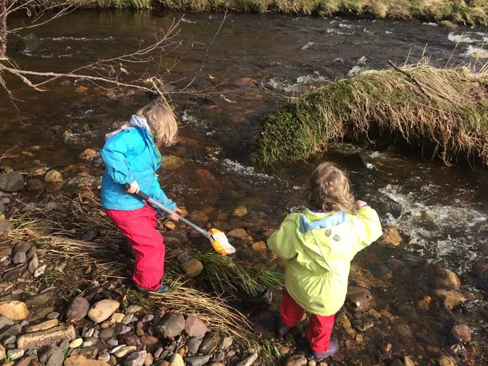 Two young children with nets paddling in a shallow stream amongst rocks and moss-covered banks.