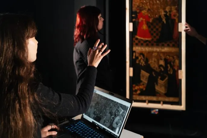 Two women examine an ornate historical painting on display, one holding a laptop showing details of the artwork.