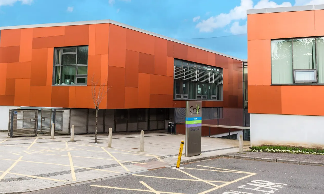 Modern orange-clad leisure centre building with glass frontage and entrance signage on a paved forecourt under blue sky.