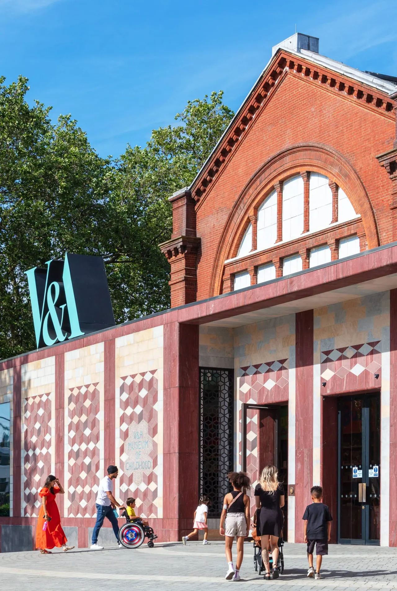 Visitors gather outside a red-brick museum building with a large arched window on a sunny day.