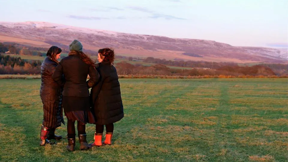 Three people standing together in a rural valley at dusk, facing distant hills.