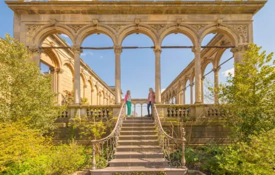 Two visitors walking through a grand stone colonnade with arched openings at a historic garden estate.