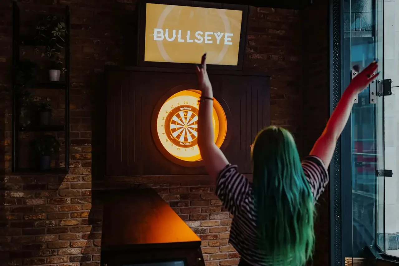Person with arm raised preparing to throw a dart at an illuminated dartboard marked "Bullseye" on a brick wall.
