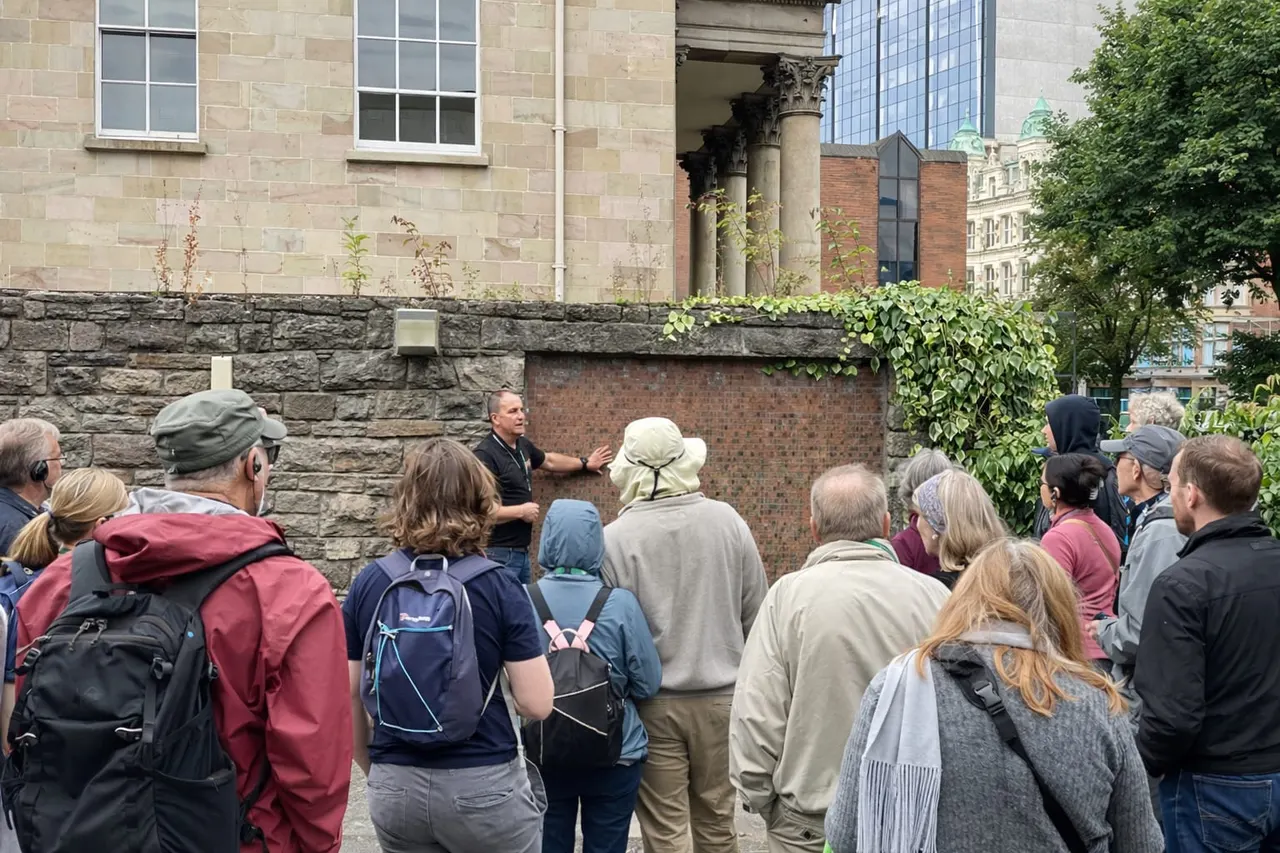 A tour guide addresses a group of visitors gathered outside a historic stone building in an urban setting.