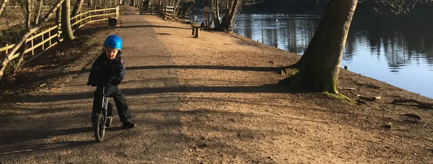 Child in blue helmet riding bike on dirt path beside river and bare trees.
