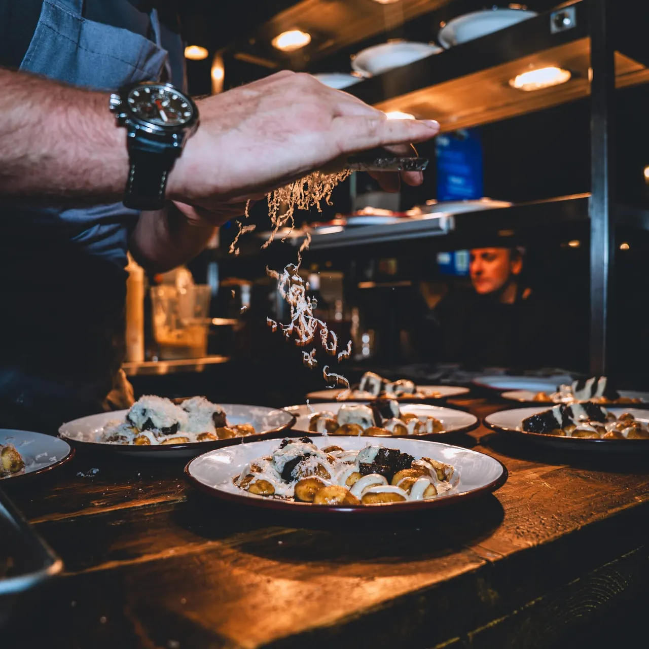 Chef sprinkles garnish over plated seafood dishes in an open kitchen at a restaurant.