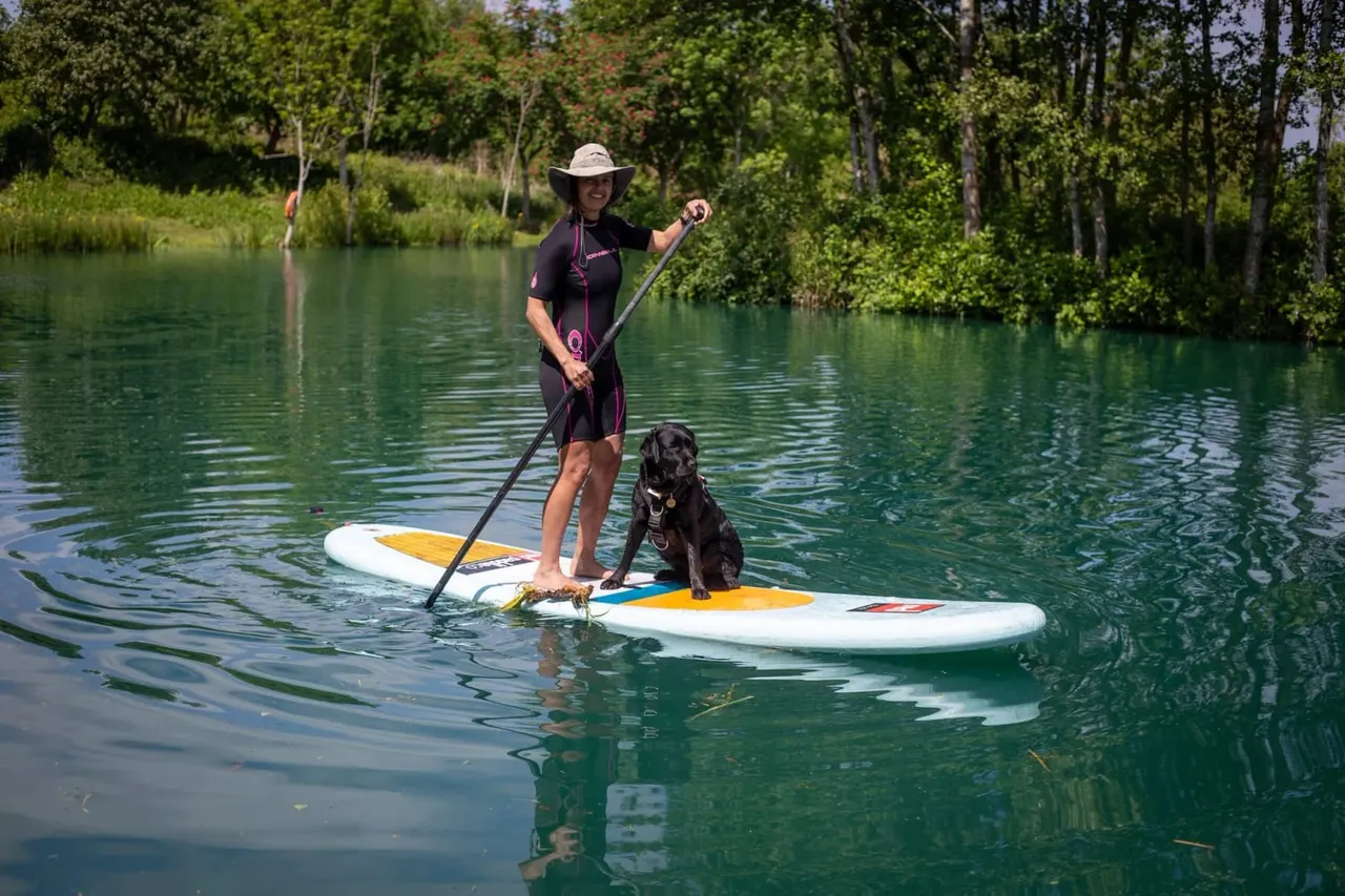 Woman paddle boarding on a calm river with a dog standing on her board, surrounded by forested banks.