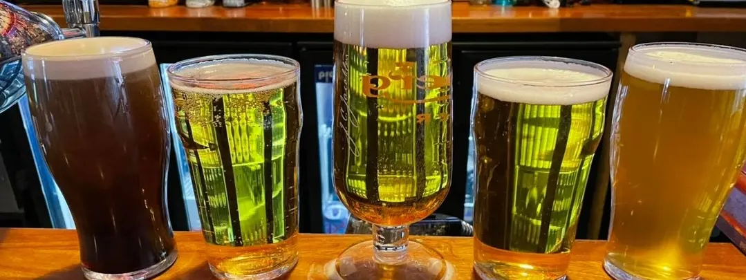 Five glasses of beer in varying shades from dark brown to pale gold lined up on a wooden bar counter.