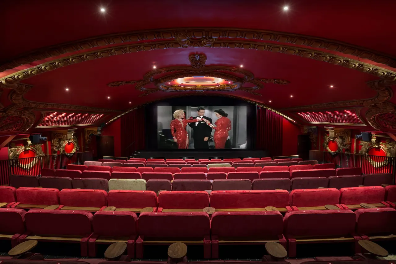 Empty cinema auditorium with red velvet seats facing a screen showing a vintage film scene.