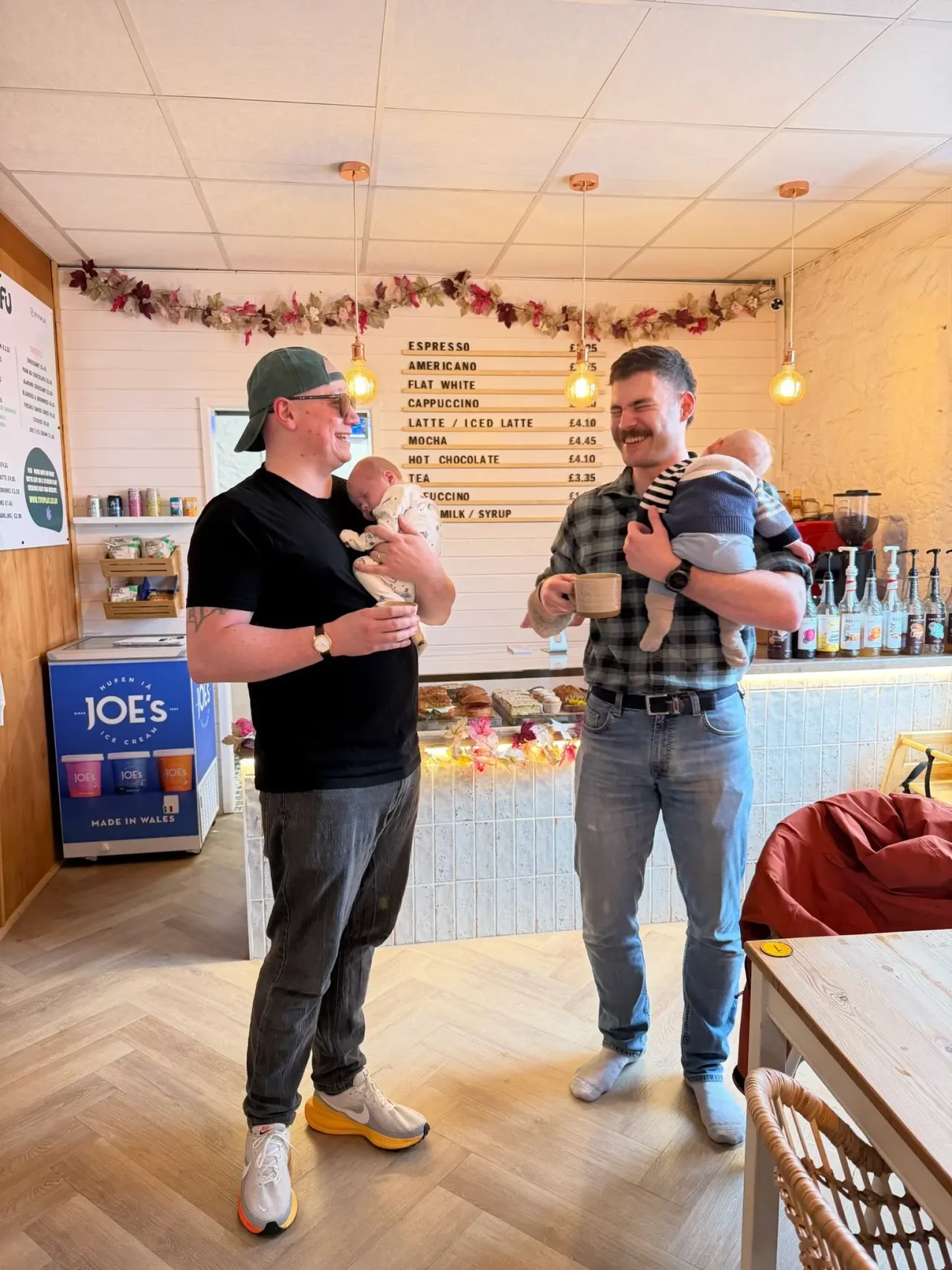 Two men holding infants chat inside a modern café decorated with floral garlands and a menu board.