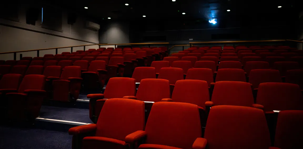 Empty cinema auditorium with rows of red velvet seats and overhead lighting.