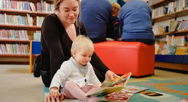 Woman and toddler looking at a picture book together on the floor of a library with bookshelves and other visitors in the background.