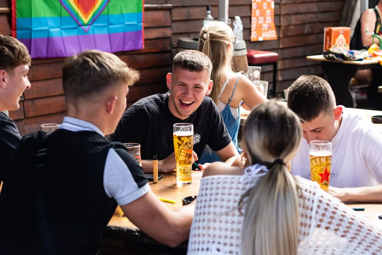 A group of young people socialise at a table with drinks in a brick-walled venue decorated with a rainbow flag.