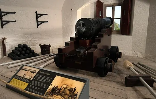 Historic cannon on wooden carriage with cannonballs and interpretive display in a whitewashed castle room.