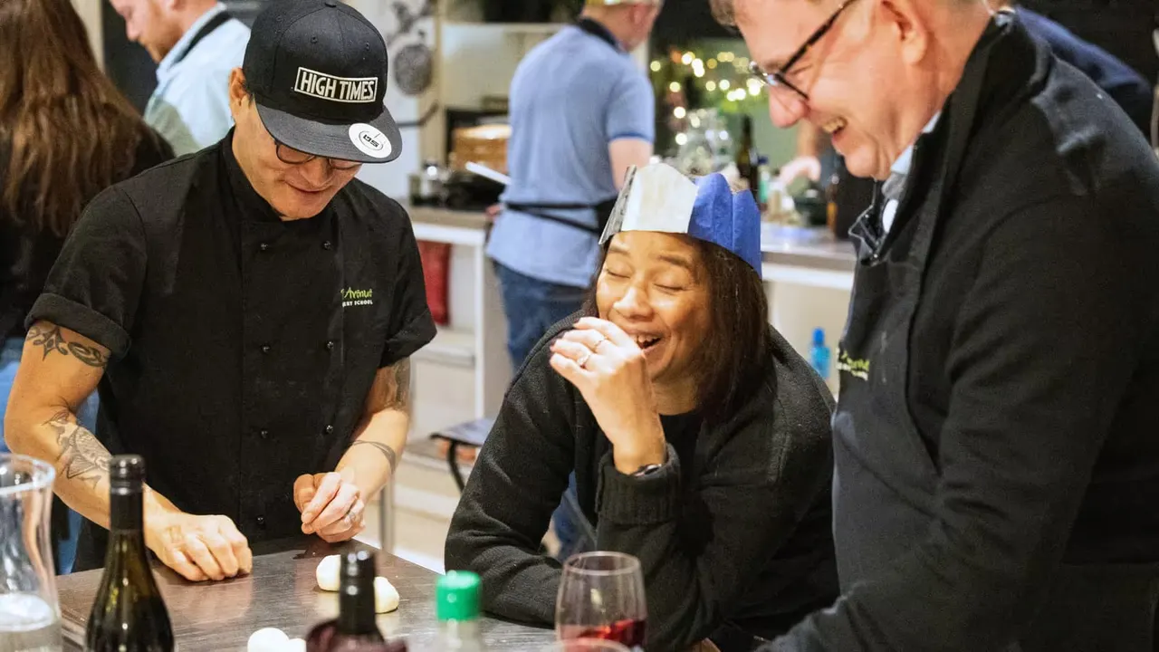 A chef in a black jacket and cap serves food to a smiling woman wearing a blue and white paper hat at a cooking class event.