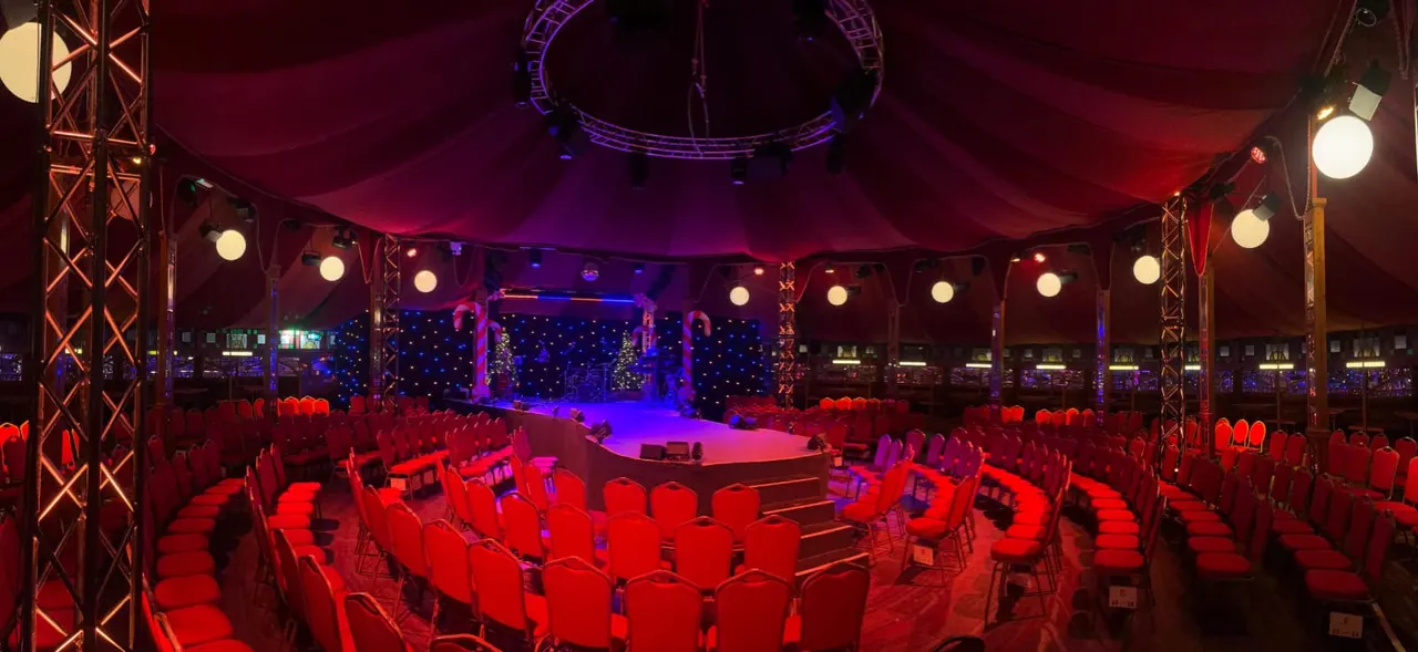 Illuminated circus-style tent with red seating, stage, and purple and blue lighting at night.