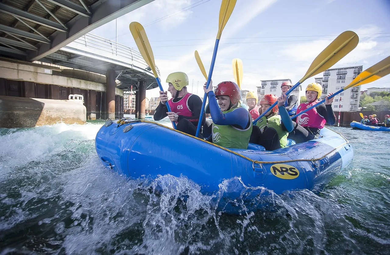 Group of people whitewater rafting in blue inflatable boats on urban river rapids beneath a bridge.