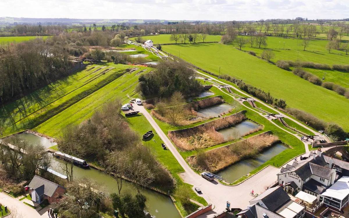 Aerial view of canal locks with moored narrowboats, green fields, and surrounding countryside in spring.