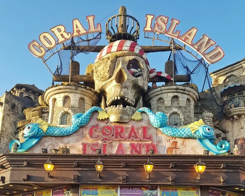 Ornate fairground attraction facade featuring a large skull with pirate-themed decoration and "Coral Island" signage against clear blue sky.