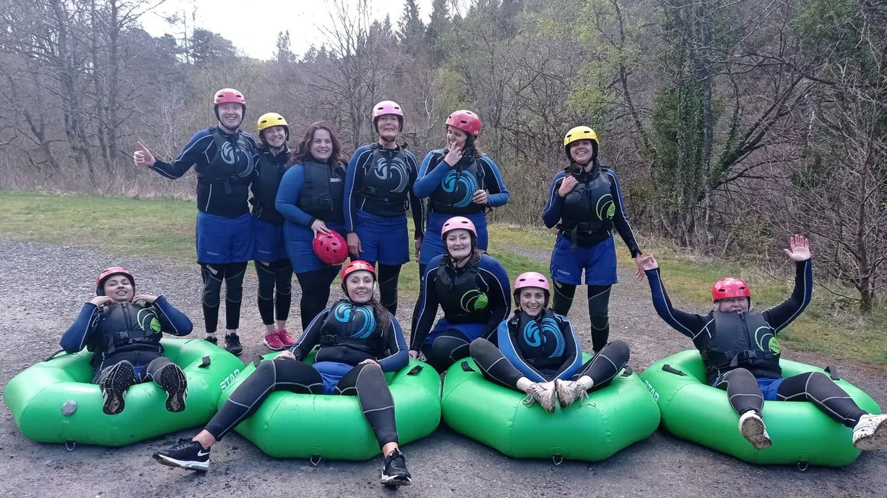 Group of people wearing helmets and wetsuits posing with green inflatable tubing by a woodland path.