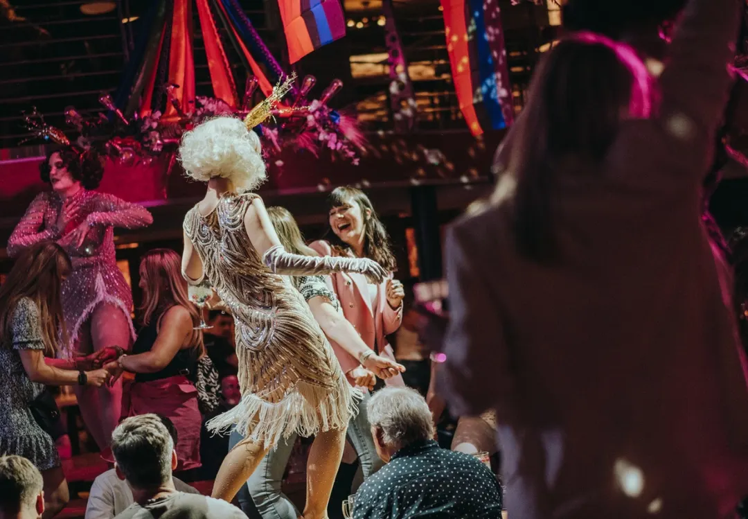 Performer in elaborate feathered costume with white wig entertaining audience at vibrant indoor venue with coloured ribbons and festive decorations.