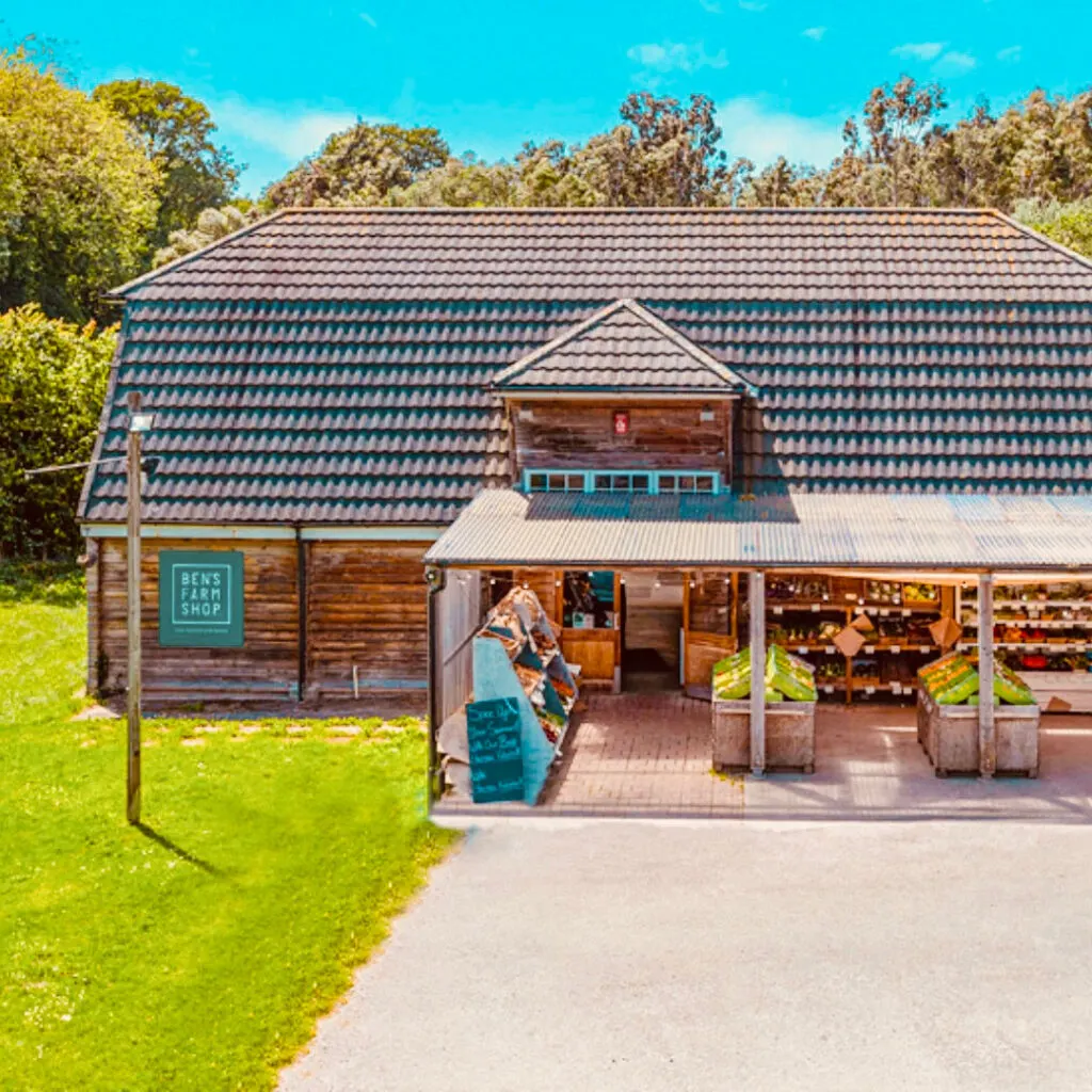 Wooden farm shop building with teal roof surrounded by green fields and trees.
