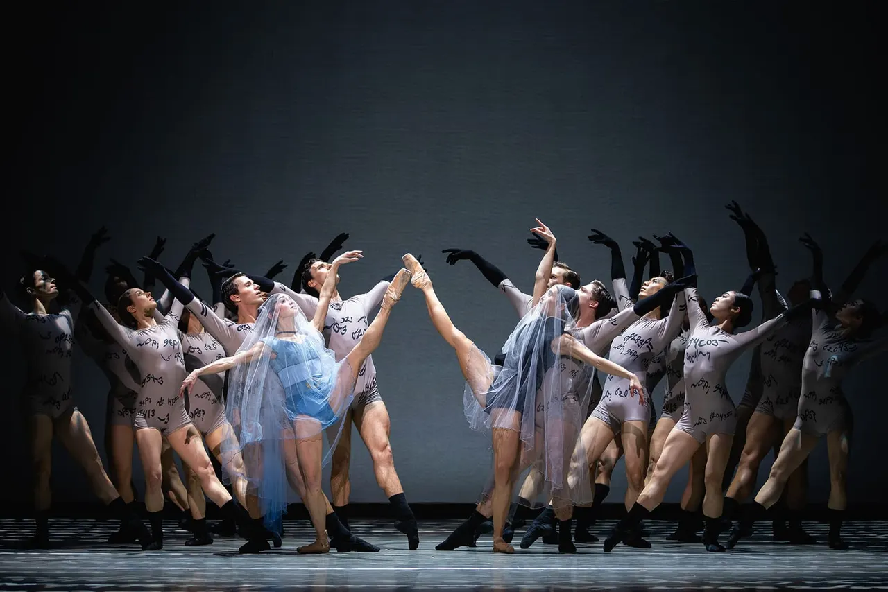 Large ensemble of ballet dancers in white and blue costumes performing synchronized movements on stage against a dark background.
