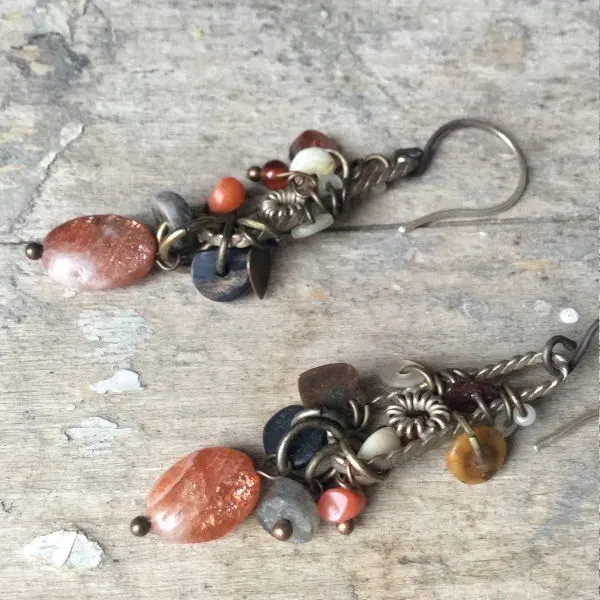 A pair of handmade beaded earrings with bronze-toned metalwork and semi-precious stone pendants on a weathered concrete surface.