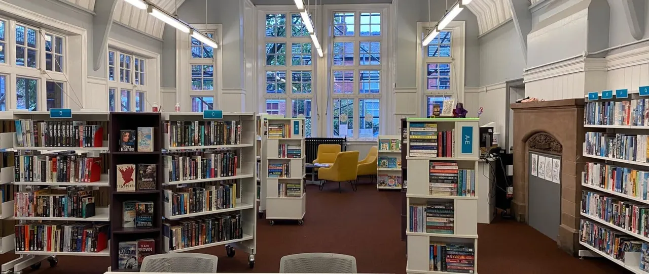 Modern library interior with bookshelves, a yellow armchair, large windows and natural lighting.