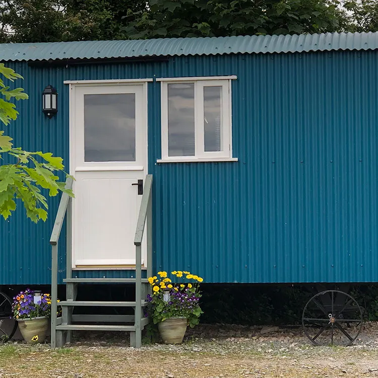 White door and window on a blue corrugated metal farm building, with flowering planters and wagon wheel nearby.