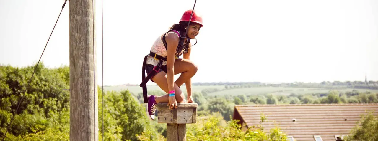 Person in safety harness and red helmet zip-lining past wooden pole with rural countryside and buildings visible below.
