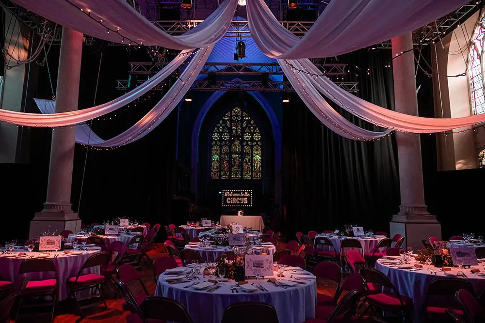 Elegant event space with round tables set for dinner under dramatic pink draped fabric and blue lighting, featuring ornate stained glass window backdrop.