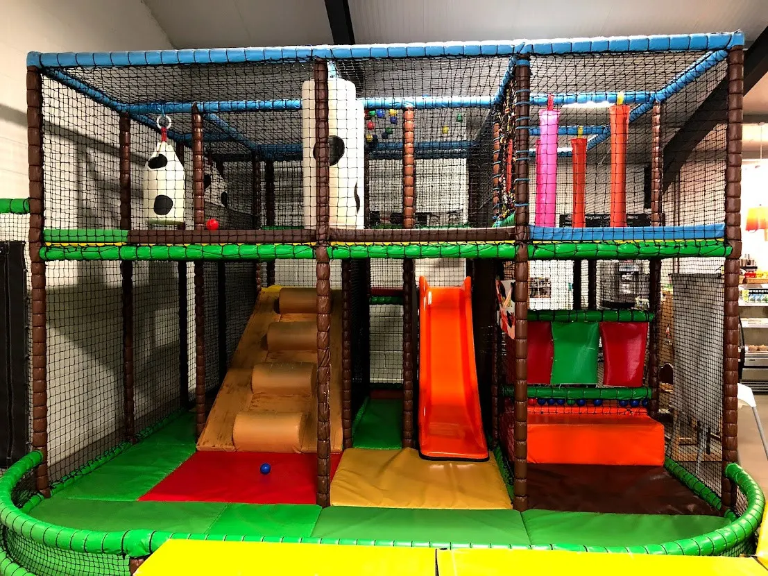 Large indoor play structure with slides, climbing elements and coloured foam padding inside a netted enclosure.