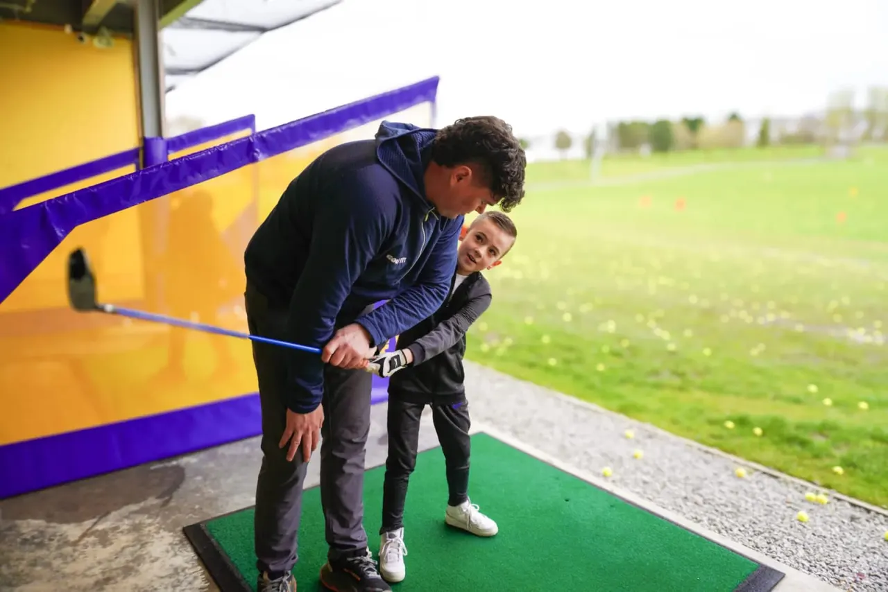 An adult and child playing mini golf together on a green course, with a colourful yellow and purple obstacle structure behind them and an open field visible in the background.