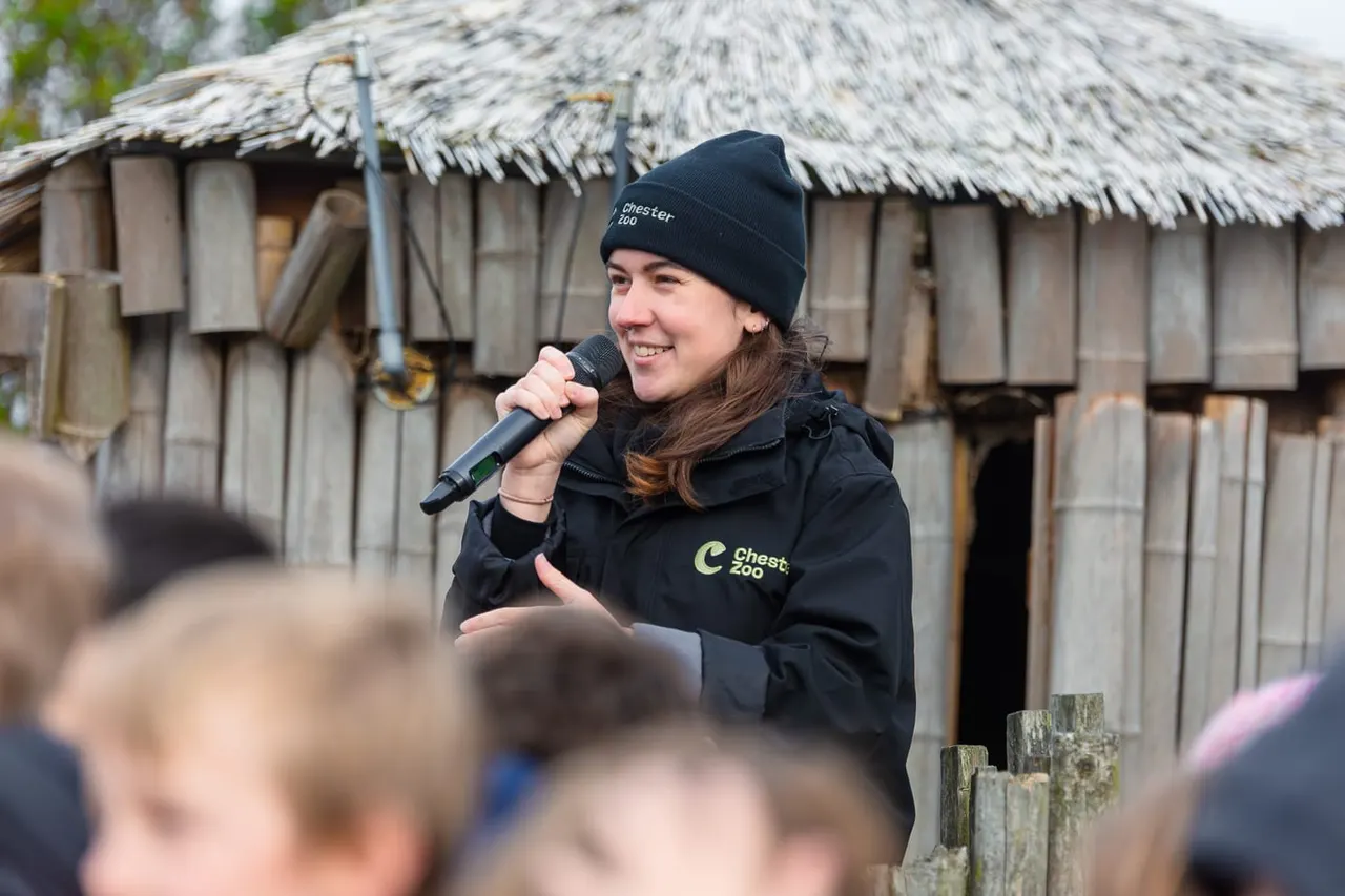 Zoo staff member speaking to visitors with microphone in front of a thatched-roof enclosure.