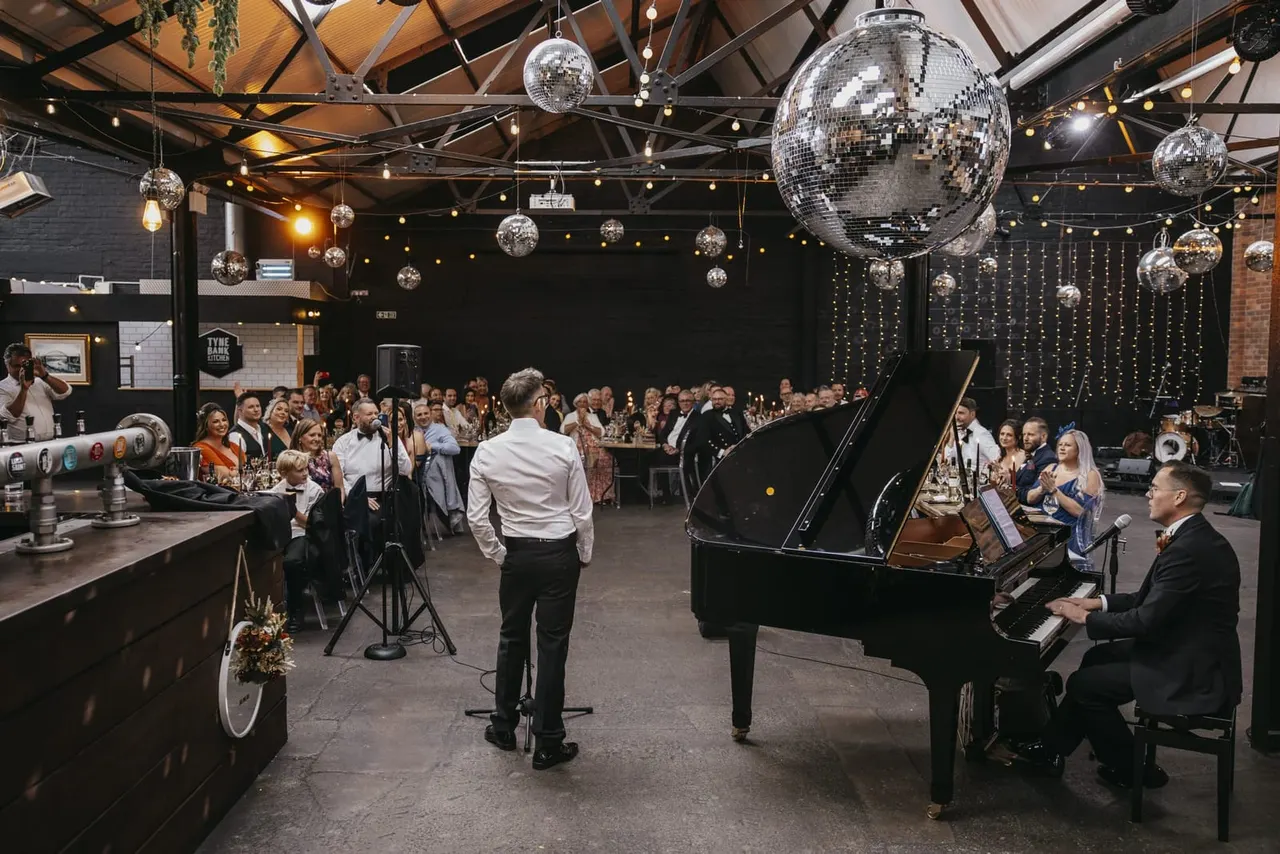 A pianist performs at a grand piano during a formal event in an industrial venue decorated with large mirror balls and string lights, with guests mingling throughout the space.