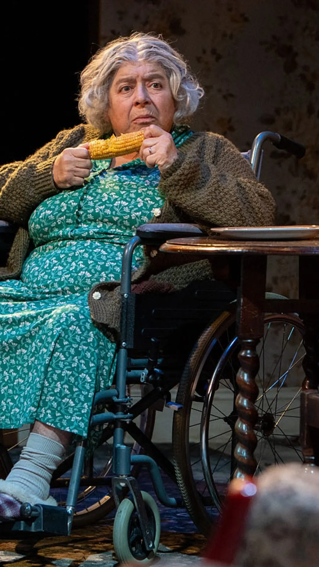 Elderly woman in wheelchair holding corn on stage during theatrical performance.