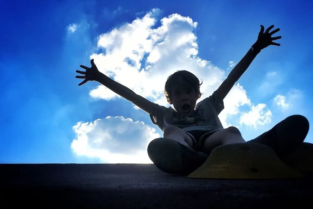 Child with arms raised in celebration against blue sky with white clouds.