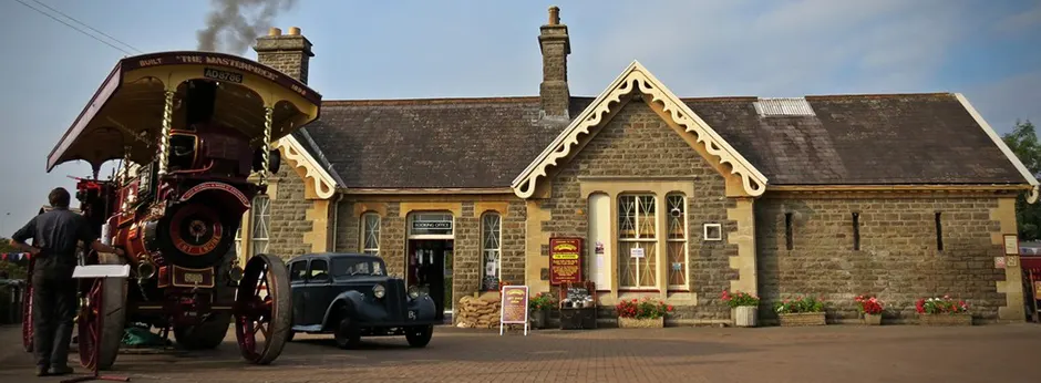 Steam locomotive and vintage vehicles parked outside a stone railway station building with cream trim.