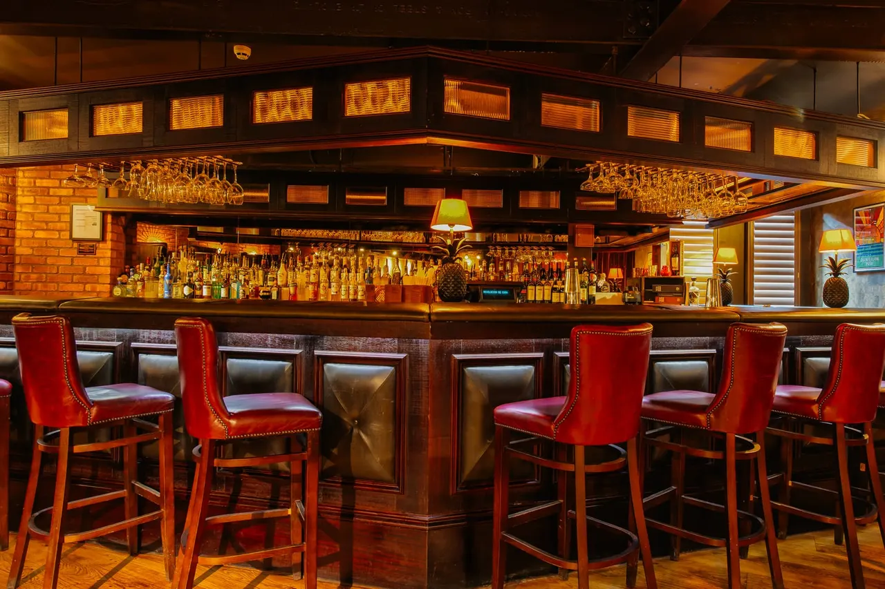 Bar counter with red upholstered stools, extensive bottle display, and warm amber lighting in an upscale cocktail bar.