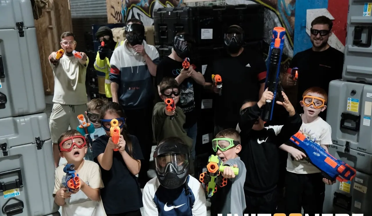 Group of children and adults wearing protective masks and goggles, holding foam dart blasters indoors at an airsoft arena.