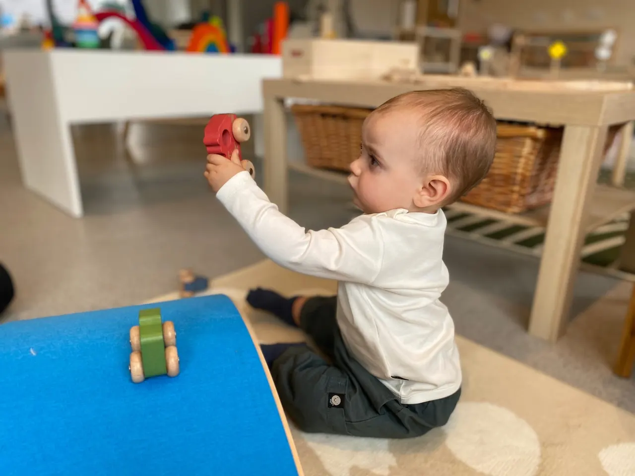 Toddler playing with wooden toys on a blue mat in a bright indoor play space.