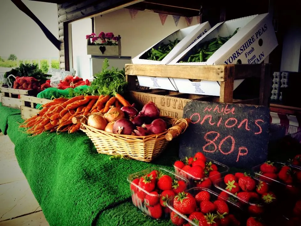 Farm shop display with fresh produce including carrots, onions, strawberries, peppers and flowers on green table.