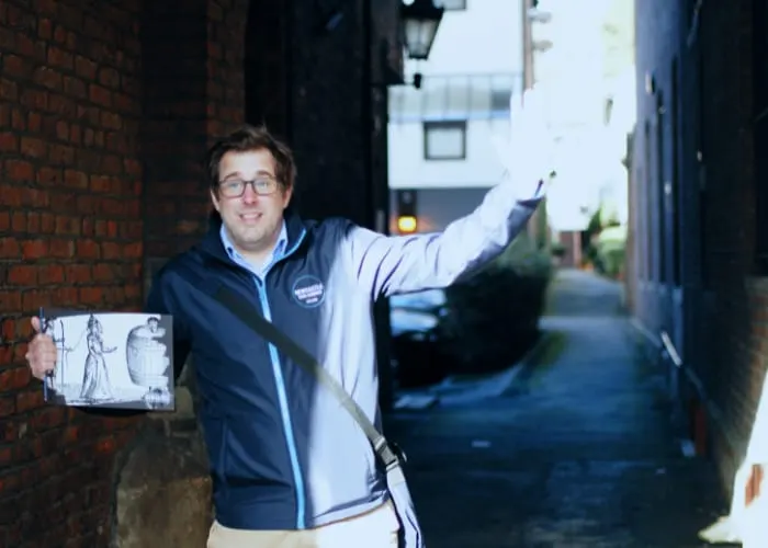 Man in glasses and jacket holding a printed guide whilst pointing down an alleyway between brick buildings.