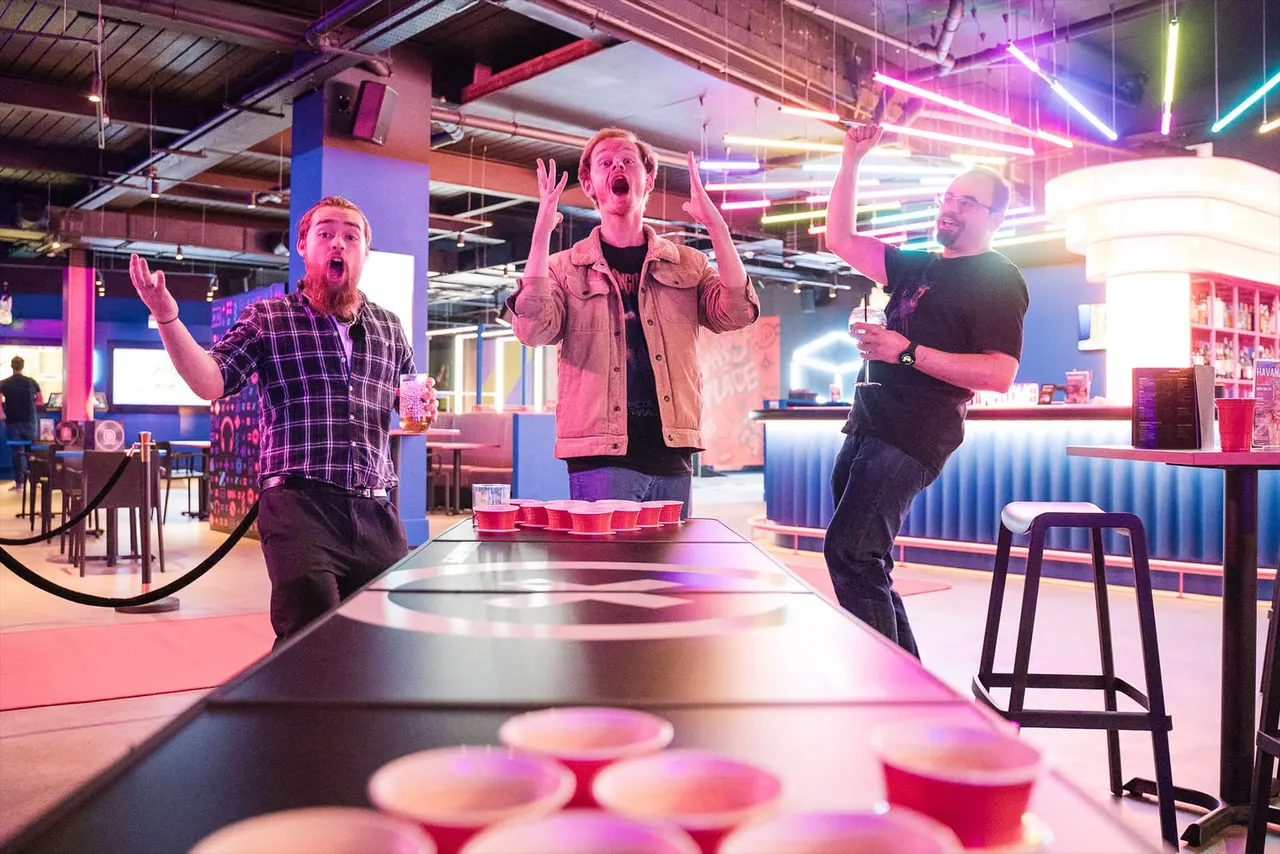 Three men playing beer pong at a neon-lit indoor entertainment venue with colourful lighting and modern furnishings.