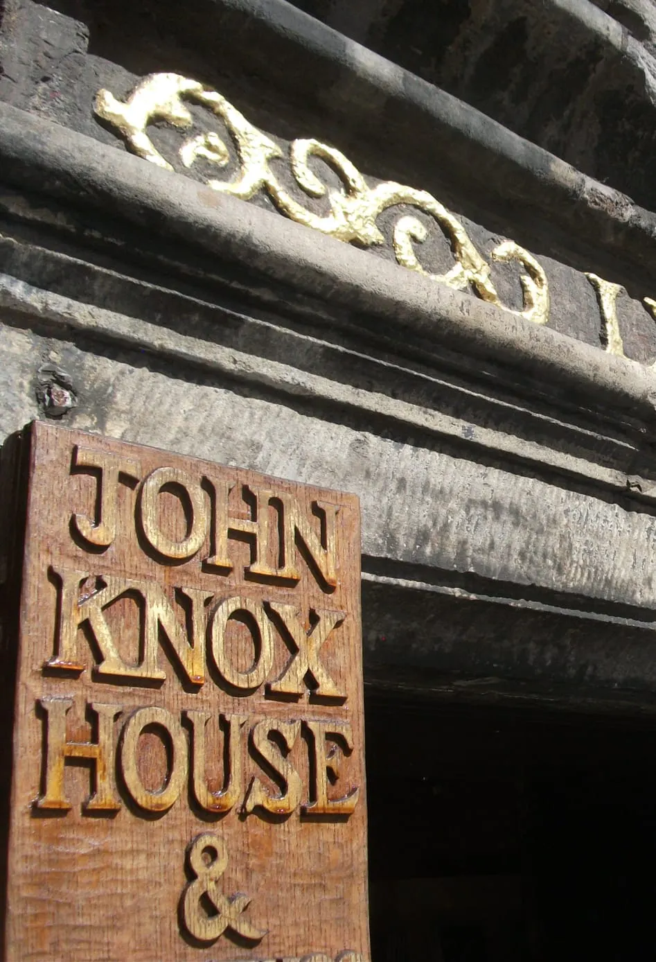 Historic stone building facade with carved golden lettering on dark stonework above a wooden sign reading "John Knox House &" mounted on the dark timber door frame.